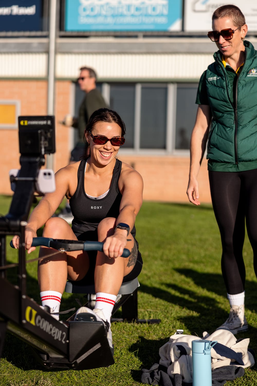 A rower smiling mid-stroke on a Concept2 erg with a coach beside her at an outdoor Row Nation session
