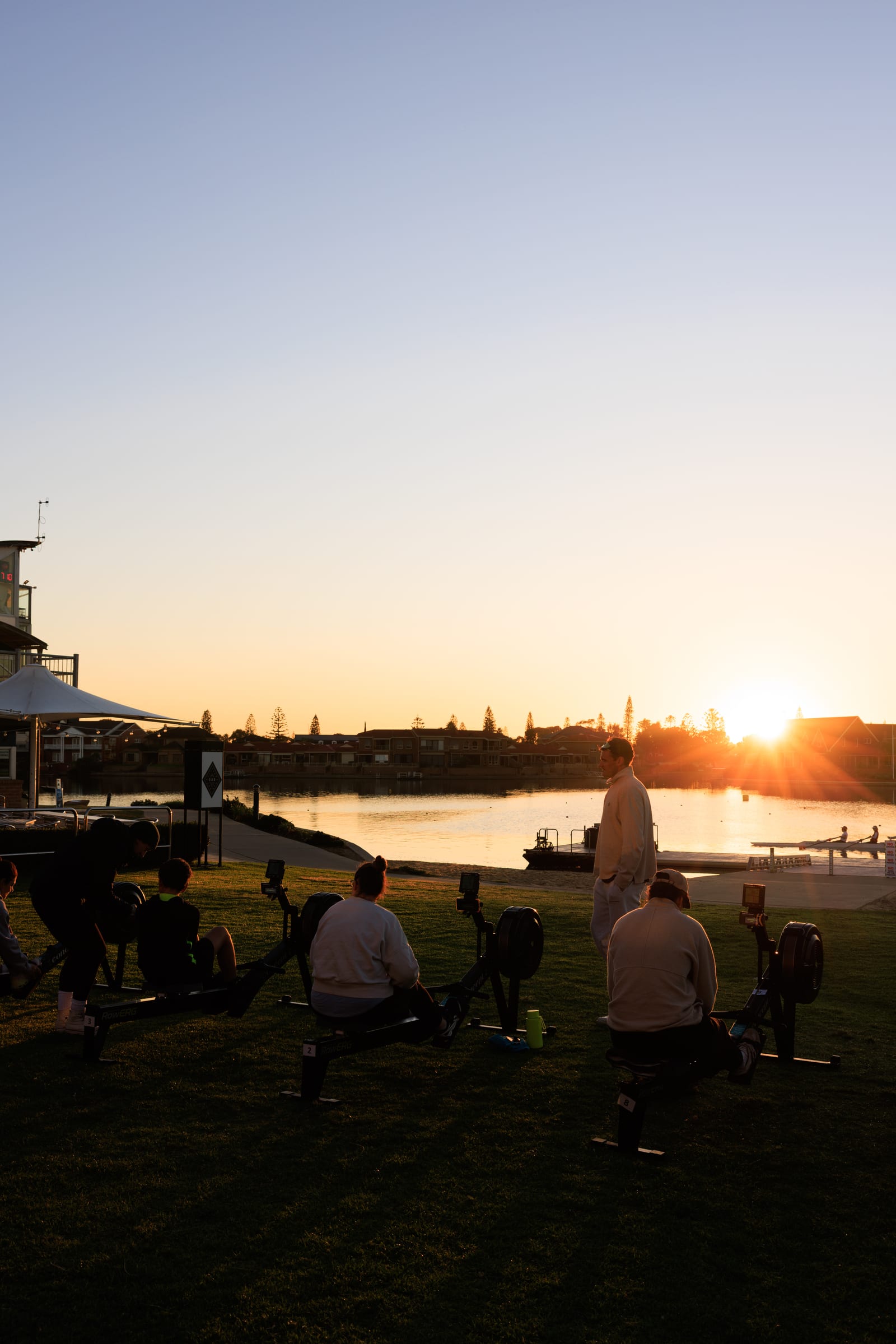 A group session on Concept2 RowErgs at sunset by the water, rowers silhouetted against the sky